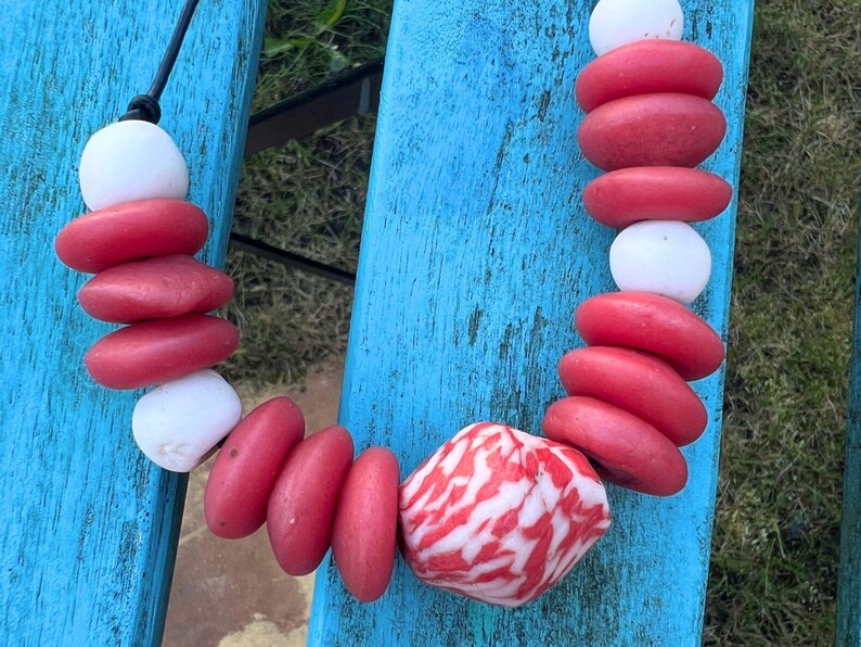 A necklace made with red and white stone beads. The necklace has a black cord and is made up of red, oval-shaped beads and white, round beads. A larger, patterned stone is at the centre.