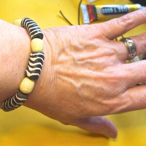 May include: A black and white striped bracelet with cream-colored beads is worn on a wrist. The bracelet's design is reminiscent of zebra stripes. A silver ring with small circular details is on a finger.