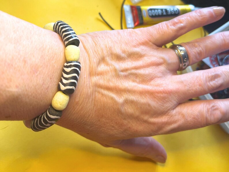 May include: A black and white striped bracelet with cream-colored beads is worn on a wrist. The bracelet's design is reminiscent of zebra stripes. A silver ring with small circular details is on a finger.
