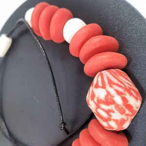 A necklace with red and white beads on a black cord. The necklace features a mix of round and oblong beads, with a larger, patterned bead at the centre. The beads are arranged on a black cord, creating a striking contrast.