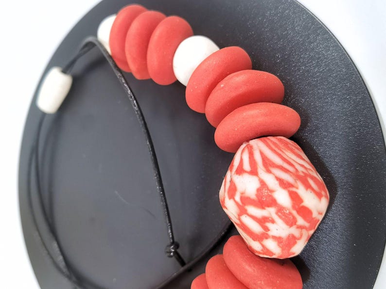 A necklace with red and white beads on a black cord. The necklace features a mix of round and oblong beads, with a larger, patterned bead at the centre. The beads are arranged on a black cord, creating a striking contrast.