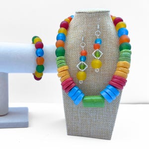 May include: A colorful beaded jewelry set featuring a necklace, earrings, and bracelet. The necklace and bracelet have a variety of round and disc-shaped beads in red, orange, yellow, green, and blue. The earrings have yellow and green beads.