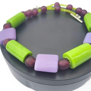 Puede incluir: Un collar colorido con cuentas grandes y brillantes. El dise&ntilde;o incluye cuentas cil&iacute;ndricas de color verde brillante, cuentas cuadradas de color lavanda y cuentas redondas m&aacute;s peque&ntilde;as de color p&uacute;rpura oscuro. El collar est&aacute; ensartado en una delicada cadena con un cierre plateado.