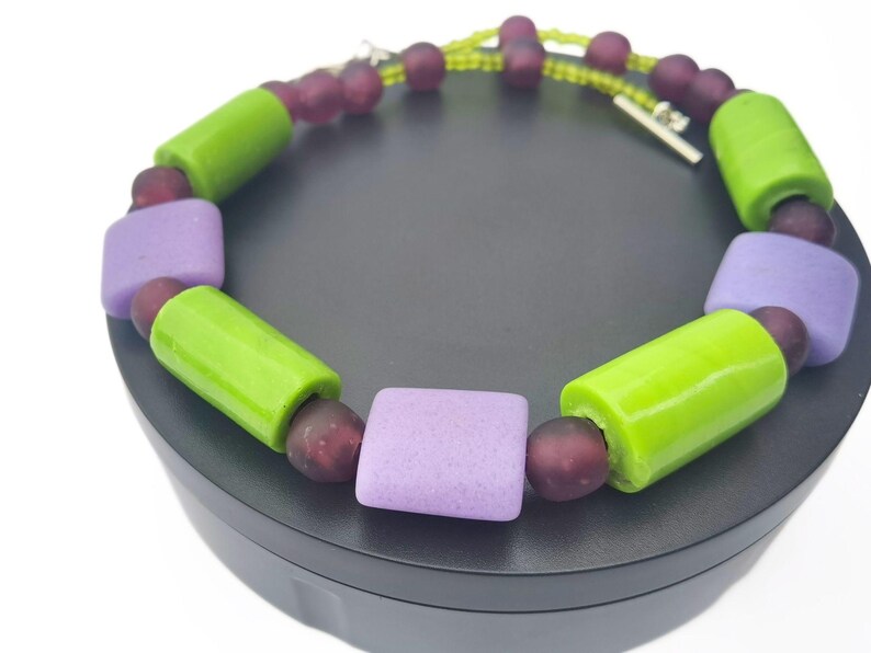 Puede incluir: Un collar colorido con cuentas grandes y brillantes. El dise&ntilde;o incluye cuentas cil&iacute;ndricas de color verde brillante, cuentas cuadradas de color lavanda y cuentas redondas m&aacute;s peque&ntilde;as de color p&uacute;rpura oscuro. El collar est&aacute; ensartado en una delicada cadena con un cierre plateado.