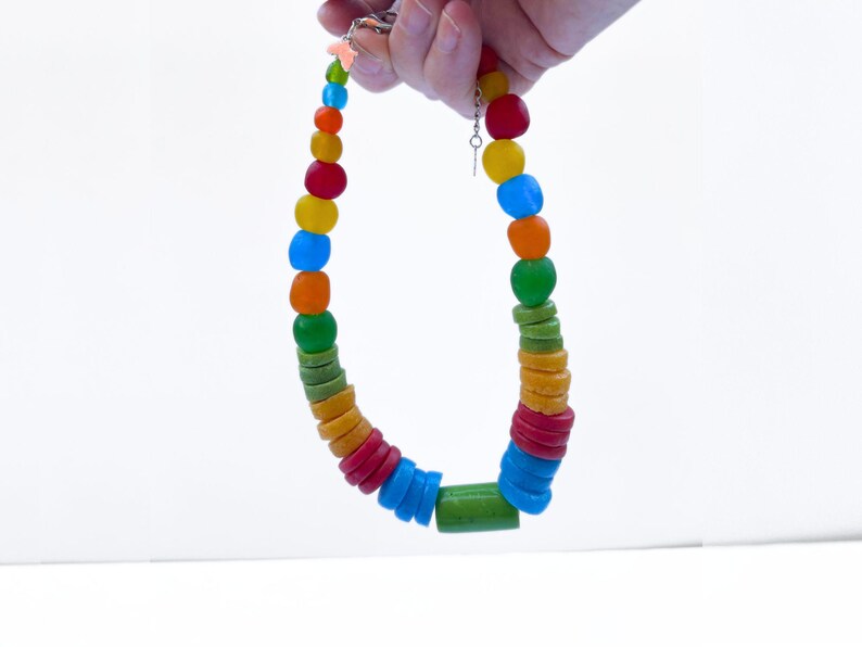 A vibrant necklace featuring a mix of round and cylindrical recycled glass beads in a rainbow of colours. The necklace is held up against a white backdrop.