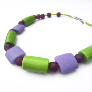 Puede incluir: Un collar colorido con cuentas cil&iacute;ndricas verdes y lavanda alternas, intercaladas con peque&ntilde;as cuentas redondas de color p&uacute;rpura oscuro. El collar est&aacute; ensartado en un cord&oacute;n verde y tiene un cierre plateado.