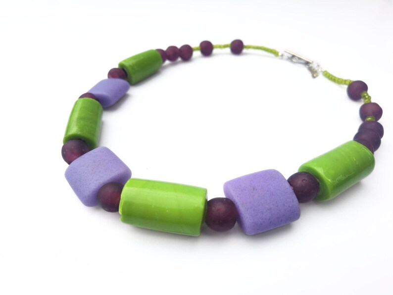 Puede incluir: Un collar colorido con cuentas cil&iacute;ndricas verdes y lavanda alternas, intercaladas con peque&ntilde;as cuentas redondas de color p&uacute;rpura oscuro. El collar est&aacute; ensartado en un cord&oacute;n verde y tiene un cierre plateado.