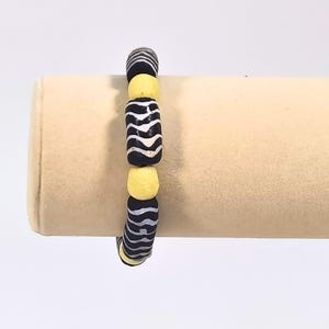 May include: A beaded bracelet featuring alternating black and white striped beads and yellow beads. The bracelet is displayed on a beige stand, showcasing its design and color contrast. This accessory is suitable for everyday wear.