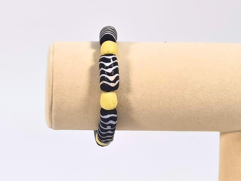May include: A beaded bracelet featuring alternating black and white striped beads and yellow beads. The bracelet is displayed on a beige stand, showcasing its design and color contrast. This accessory is suitable for everyday wear.
