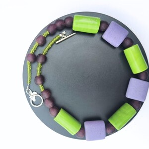 Puede incluir: Un collar con una mezcla de cuentas moradas, verdes y lavanda. El collar tiene un cierre plateado y se muestra sobre una superficie circular gris oscuro sobre un fondo blanco. Las cuentas son de varias formas y tama&ntilde;os.