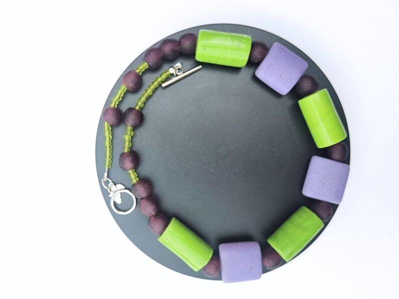 Puede incluir: Un collar con una mezcla de cuentas moradas, verdes y lavanda. El collar tiene un cierre plateado y se muestra sobre una superficie circular gris oscuro sobre un fondo blanco. Las cuentas son de varias formas y tama&ntilde;os.