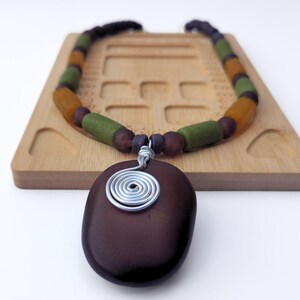 Peut inclure: Un collier artisanal avec un pendentif ovale marron fonc&eacute; orn&eacute; d'un motif en spirale en fil d'argent. Le collier est compos&eacute; de perles marron, vertes et ambr&eacute;es. Le collier est pr&eacute;sent&eacute; sur un plateau en bois.