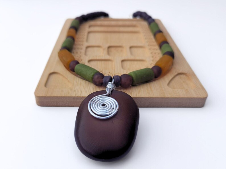 Peut inclure: Un collier artisanal avec un pendentif ovale marron fonc&eacute; orn&eacute; d'un motif en spirale en fil d'argent. Le collier est compos&eacute; de perles marron, vertes et ambr&eacute;es. Le collier est pr&eacute;sent&eacute; sur un plateau en bois.