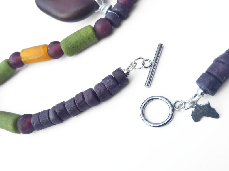 Peut inclure: Gros plan sur un collier fait &agrave; la main avec des perles color&eacute;es. Le collier comprend des perles violettes, vertes et jaunes, un fermoir argent&eacute; et un charm en forme de continent africain.