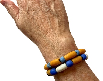 Puede incluir: Una pulsera de varias vueltas con cuentas cilíndricas y esféricas en tonos amarillo mostaza, azul claro, azul real y blanco roto. Las cuentas están ensartadas en un patrón repetitivo, creando una apariencia texturizada y superpuesta.