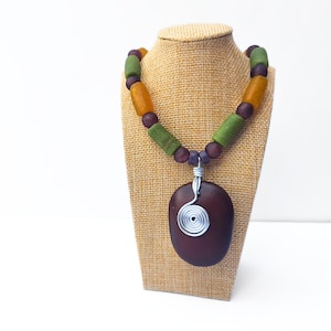 Peut inclure: Un collier artisanal avec un pendentif ovale marron fonc&eacute; et une spirale en fil d'argent. Le collier a un fil de perles avec des perles vertes, orange et violettes. Le collier est pr&eacute;sent&eacute; sur un pr&eacute;sentoir &agrave; bijoux beige.