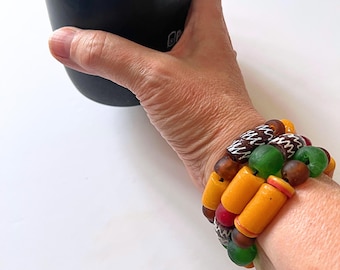 Puede incluir: Una pulsera de múltiples hilos que presenta una variedad de cuentas cilíndricas y esféricas en tonos vibrantes de amarillo, verde, marrón y rojo. Algunas cuentas muestran intrincados patrones blancos grabados, mientras otras tienen un acabado liso.