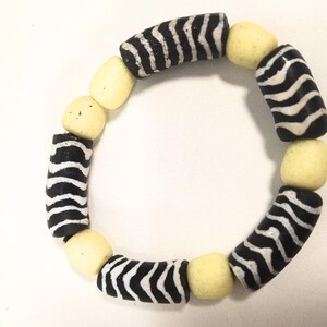 May include: A handmade bracelet featuring alternating black and white zebra-striped beads and pale yellow, rounded beads. The bracelet is circular and likely designed to be worn on the wrist. The beads are strung together to form a complete circle.