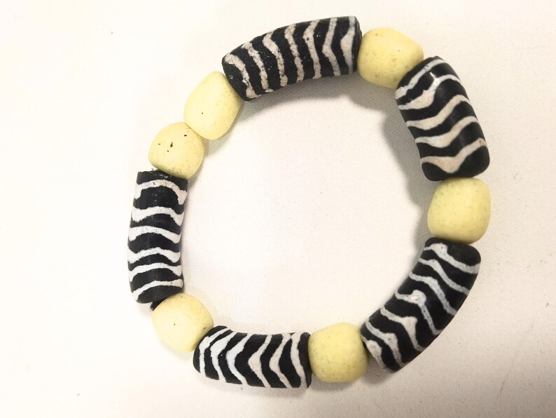 May include: A handmade bracelet featuring alternating black and white zebra-striped beads and pale yellow, rounded beads. The bracelet is circular and likely designed to be worn on the wrist. The beads are strung together to form a complete circle.