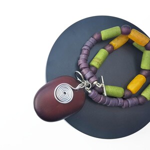 Peut inclure: Un collier artisanal avec un grand pendentif ovale rouge fonc&eacute; orn&eacute; d'un motif en spirale en fil d'argent. Le collier est compos&eacute; de perles violettes et de perles cylindriques vertes et jaunes. Le collier est pr&eacute;sent&eacute; sur une surface gris fonc&eacute;.