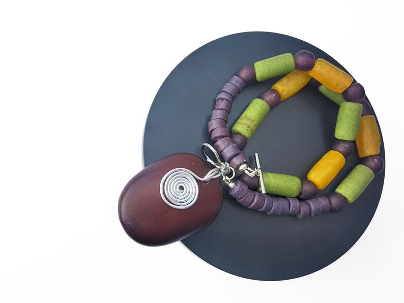 Peut inclure: Un collier artisanal avec un grand pendentif ovale rouge fonc&eacute; orn&eacute; d'un motif en spirale en fil d'argent. Le collier est compos&eacute; de perles violettes et de perles cylindriques vertes et jaunes. Le collier est pr&eacute;sent&eacute; sur une surface gris fonc&eacute;.