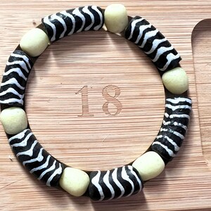 May include: A circular bracelet with black and white zebra-striped beads and cream-colored spacer beads. The bracelet is resting on a wooden surface. The number "18" is etched into the wood.