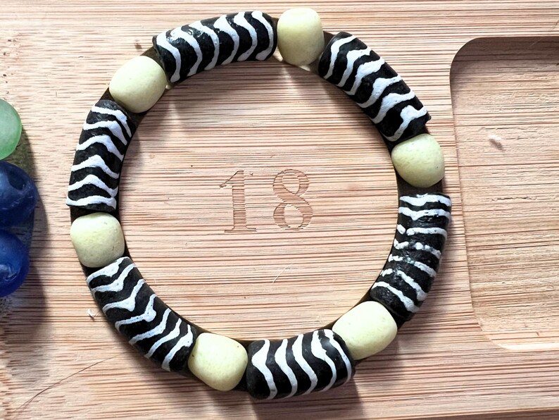May include: A circular bracelet with black and white zebra-striped beads and cream-colored spacer beads. The bracelet is resting on a wooden surface. The number "18" is etched into the wood.