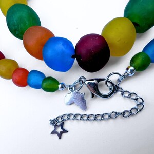 The closure of a colourful recycled glass bead necklace with large, oval beads in shades of red, orange, yellow, green, and blue. The necklace has a silver chain to adjust the length and is closed by a lobster clasp, with a star and an African charm.