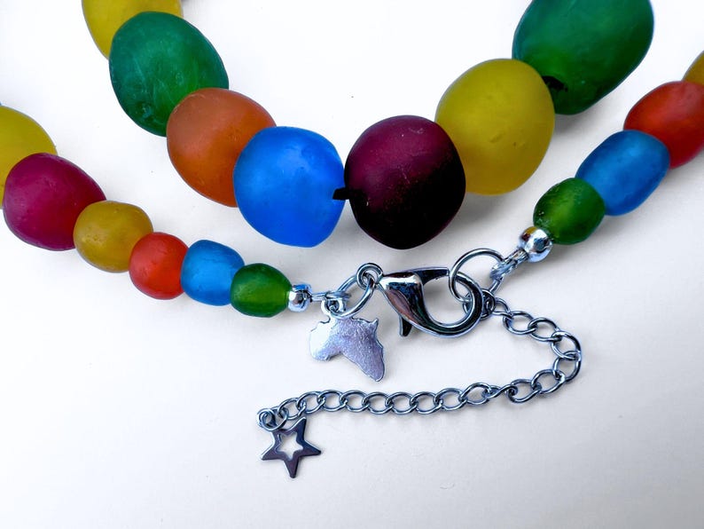 The closure of a colourful recycled glass bead necklace with large, oval beads in shades of red, orange, yellow, green, and blue. The necklace has a silver chain to adjust the length and is closed by a lobster clasp, with a star and an African charm.