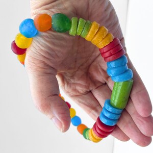 A vibrant necklace made from a mix of rainbow coloured recycled glass beads. The necklace features tubular, disc and round beads in red, yellow, blue, green, and orange. The bracelet is held in a hand clearly showing the size of the central green focal bead.