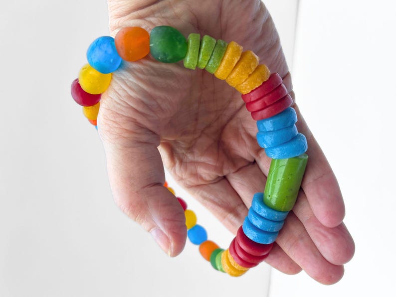 A vibrant necklace made from a mix of rainbow coloured recycled glass beads. The necklace features tubular, disc and round beads in red, yellow, blue, green, and orange. The bracelet is held in a hand clearly showing the size of the central green focal bead.