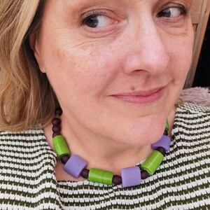 Puede incluir: Un collar colorido con cuentas rectangulares grandes en tonos verdes y lavanda, intercaladas con cuentas m&aacute;s peque&ntilde;as de color marr&oacute;n oscuro. El collar se usa sobre una blusa a rayas verdes y blancas.