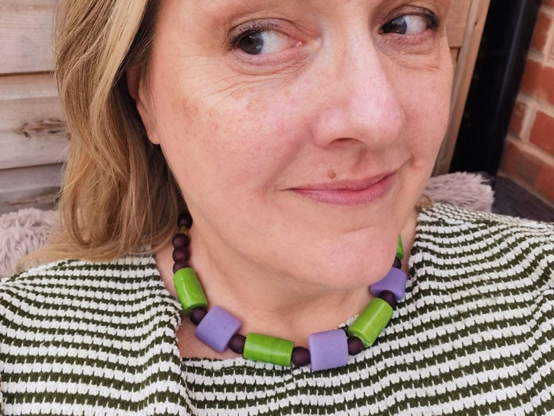 Puede incluir: Un collar colorido con cuentas rectangulares grandes en tonos verdes y lavanda, intercaladas con cuentas m&aacute;s peque&ntilde;as de color marr&oacute;n oscuro. El collar se usa sobre una blusa a rayas verdes y blancas.
