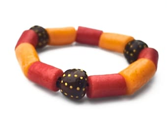 May include: A bracelet composed of cylindrical beads in alternating red and orange colors, interspersed with dark brown spherical beads featuring small yellow dots. The beads are strung together on a dark cord, forming a circular loop pattern.
