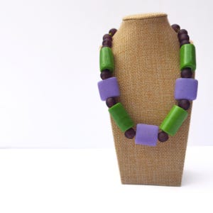 Puede incluir: Un collar colorido con cuentas verdes, moradas y marr&oacute;n oscuro. El collar presenta cuentas rectangulares y redondas en un dise&ntilde;o geom&eacute;trico audaz. El collar se exhibe en un expositor de joyer&iacute;a marr&oacute;n sobre un fondo blanco.