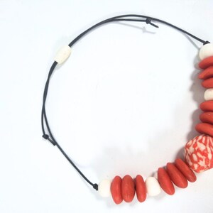 A necklace with a black cord and a sliding clasp. It features red and white beads, with a larger patterned bead with red and white swirls. The necklace is designed with a mix of shapes and colours.