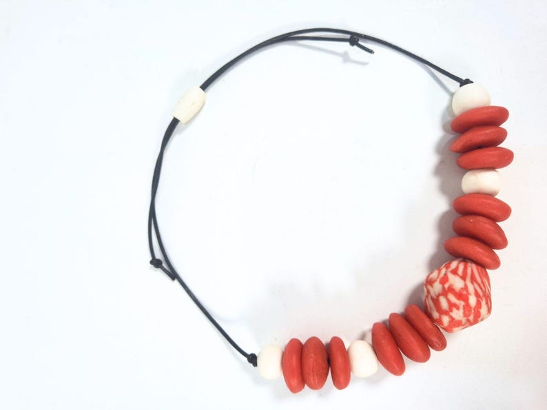 A necklace with a black cord and a sliding clasp. It features red and white beads, with a larger patterned bead with red and white swirls. The necklace is designed with a mix of shapes and colours.