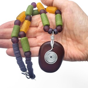 Peut inclure: Un collier artisanal avec un pendentif ovale marron fonc&eacute; et une spirale en fil d'argent. Le collier est compos&eacute; de perles violettes, vertes et jaunes. Le collier est tenu dans une main sur fond blanc.