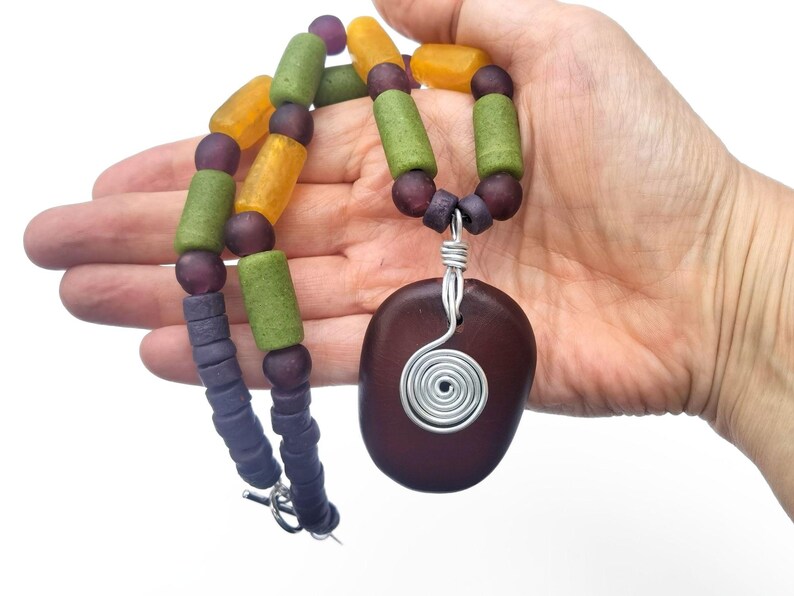 Peut inclure: Un collier artisanal avec un pendentif ovale marron fonc&eacute; et une spirale en fil d'argent. Le collier est compos&eacute; de perles violettes, vertes et jaunes. Le collier est tenu dans une main sur fond blanc.