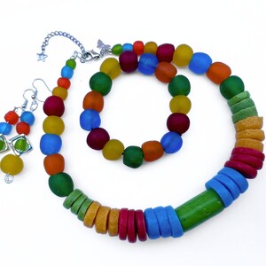 A vibrant beaded jewellery set comprising a necklace, bracelet, and earrings all made with recycled glass beads from Ghana. The necklace and bracelet feature round and disc shaped beads in shades of red, yellow, green, blue, and orange. The earrings are dangly and feature coloured beads stacked on top of each other.