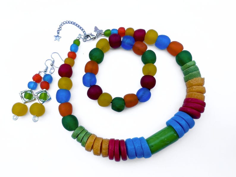A vibrant beaded jewellery set comprising a necklace, bracelet, and earrings all made with recycled glass beads from Ghana. The necklace and bracelet feature round and disc shaped beads in shades of red, yellow, green, blue, and orange. The earrings are dangly and feature coloured beads stacked on top of each other.