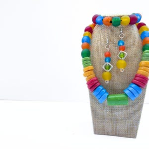 A vibrant recycled glass bead jewellery set consisting of a necklace, bracelet, and earring set. The necklace and bracelet are made of large, round beads in shades of red, orange, yellow, green, and blue. The earrings have a similar colour palette of smaller beads with geometric details.