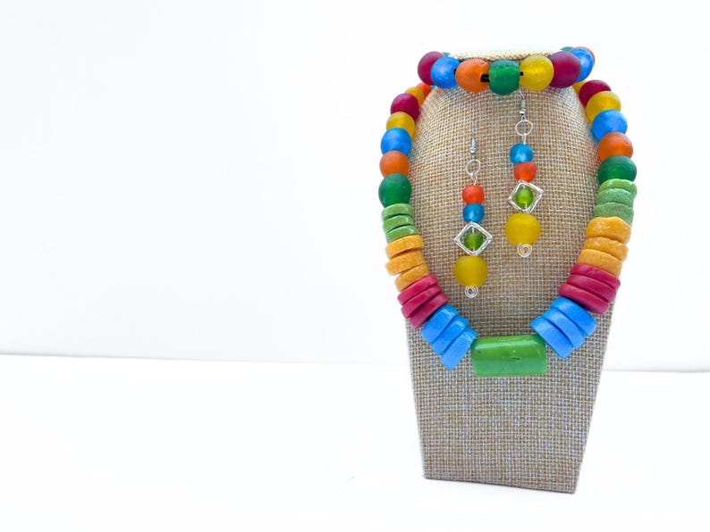 A vibrant recycled glass bead jewellery set consisting of a necklace, bracelet, and earring set. The necklace and bracelet are made of large, round beads in shades of red, orange, yellow, green, and blue. The earrings have a similar colour palette of smaller beads with geometric details.