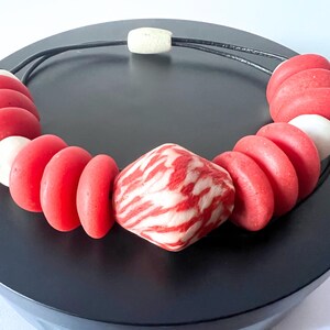 A striking necklace with large red and white beads. The necklace has a black cord and a central patterned bead. The beads are arranged in a circular pattern. The necklace is displayed on a black surface. The necklace is a statement piece.