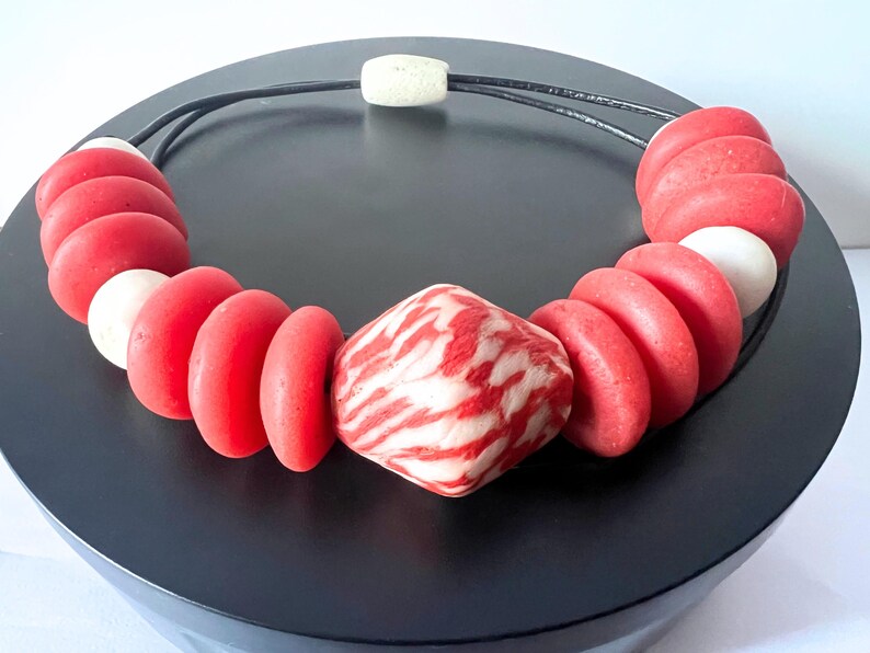 A striking necklace with large red and white beads. The necklace has a black cord and a central patterned bead. The beads are arranged in a circular pattern. The necklace is displayed on a black surface. The necklace is a statement piece.
