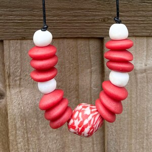 A necklace with red and white beads on a black cord. The necklace features stacked, flat, red, round beads interspersed with white, round beads. A larger, patterned bead with red and white swirls is at the centre of the necklace.