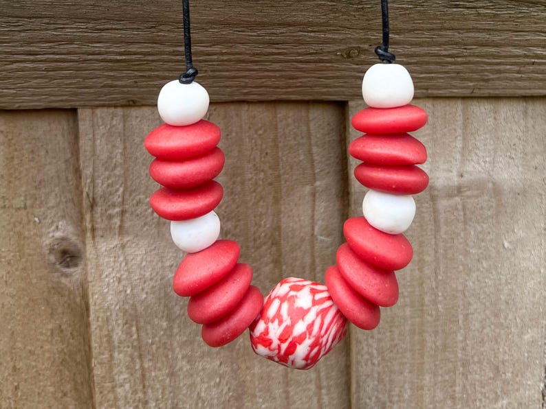 A necklace with red and white beads on a black cord. The necklace features stacked, flat, red, round beads interspersed with white, round beads. A larger, patterned bead with red and white swirls is at the centre of the necklace.
