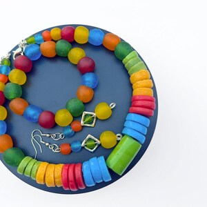 Op de afbeelding: Een kleurrijke sieradenset met een ketting, armband en oorbellen. De ketting en armband hebben ronde, matte kralen in rood, oranje, geel, groen en blauw. De oorbellen hebben vierkante groene accenten.