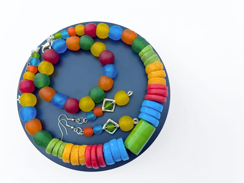 Op de afbeelding: Een kleurrijke sieradenset met een ketting, armband en oorbellen. De ketting en armband hebben ronde, matte kralen in rood, oranje, geel, groen en blauw. De oorbellen hebben vierkante groene accenten.