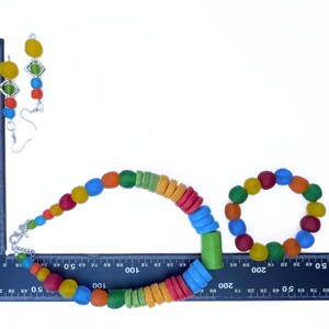 A vibrant jewellery set comprising a necklace, bracelet, and earrings. The necklace and bracelet feature round, multi-coloured beads. The earrings have yellow and green details. The jewellery is displayed on a white surface against a ruler to show the scale.